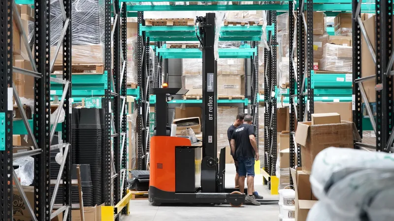 A warehouse worker guiding a forklift between tall shelves stacked with boxed inventory