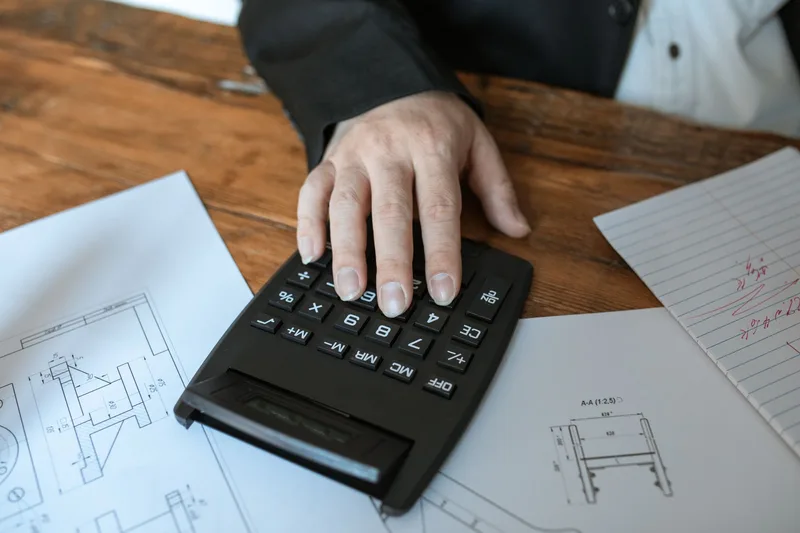 Close-up of a hand using a calculator to work through cost figures on a desk