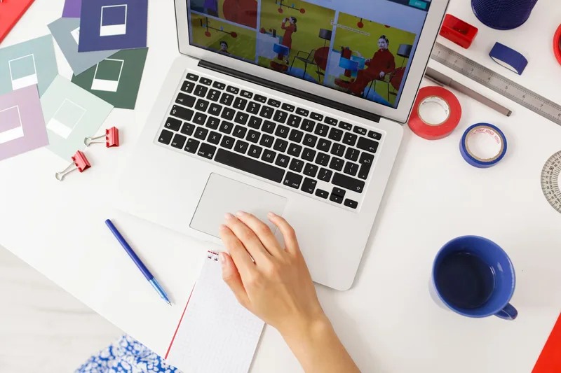 An overhead view of a laptop screen showing a colorful pin layout, surrounded by design swatches, tape, and a notebook on a creative workspace