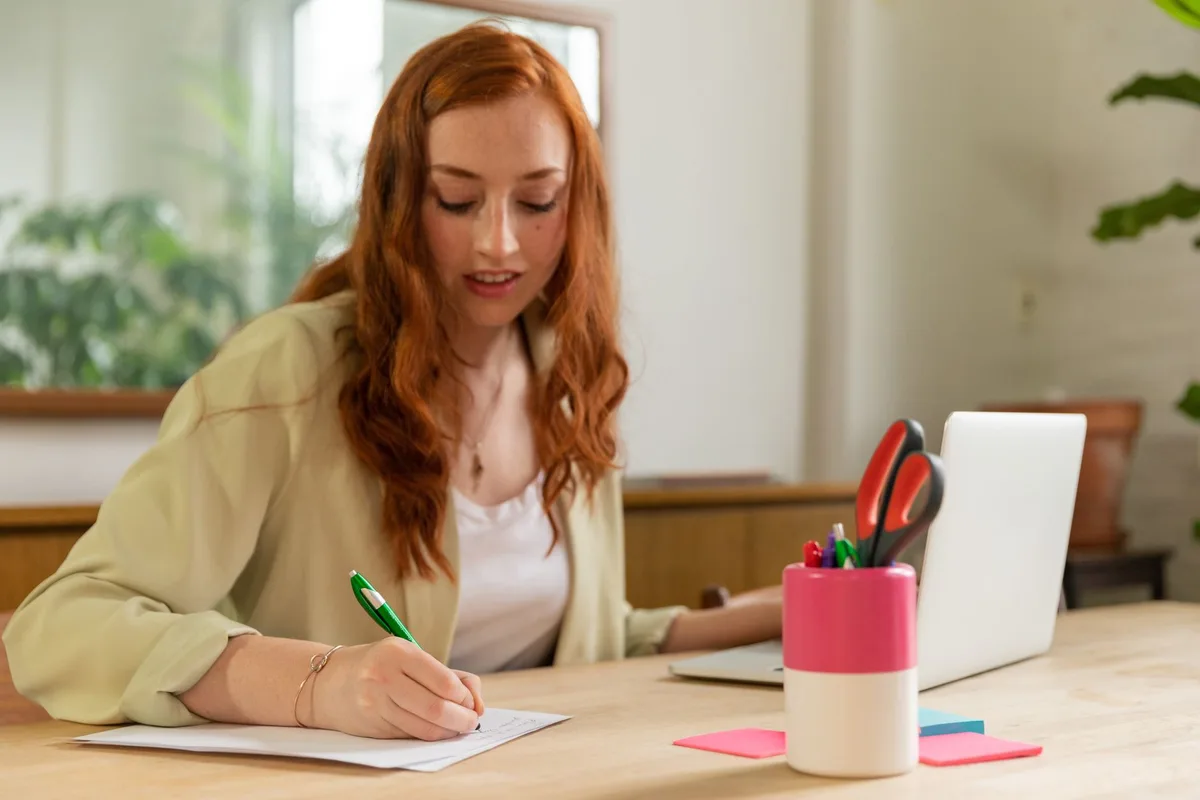 Woman writing on paper at a home office desk with a laptop, planning her virtual assistant business