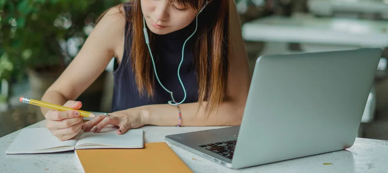 Woman wearing headphones taking notes in a notebook next to her open laptop while studying