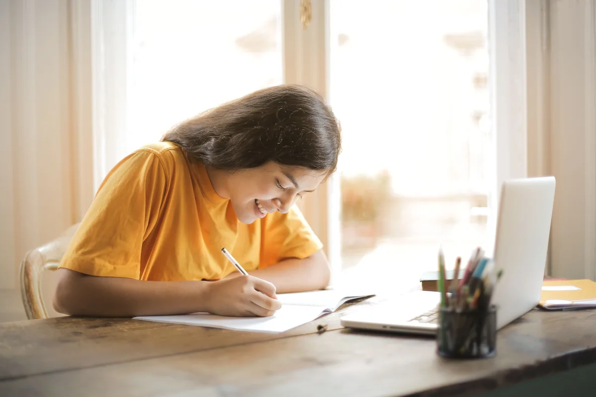Woman smiling while taking notes at a desk with an open laptop nearby, planning her virtual assistant certification path