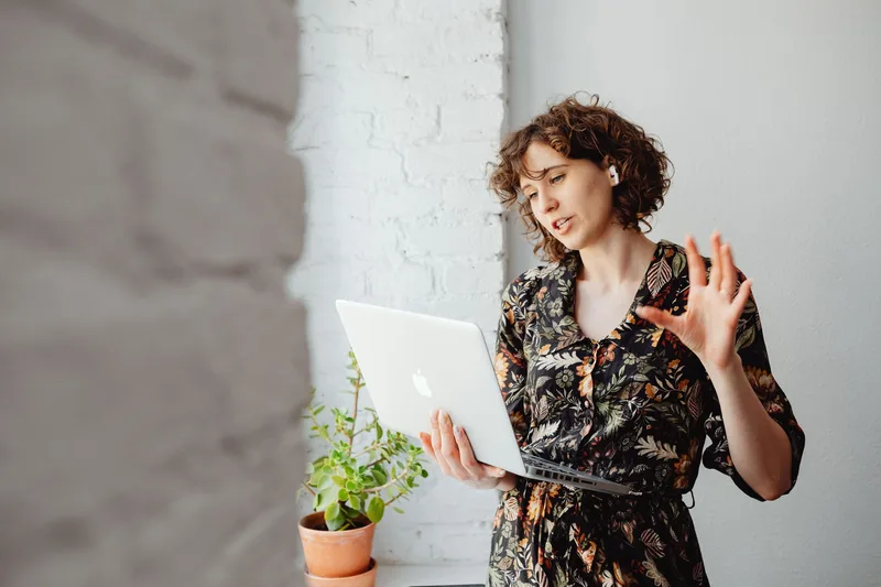 A woman with curly hair conducting an online meeting on her laptop indoors, wearing wireless headphones