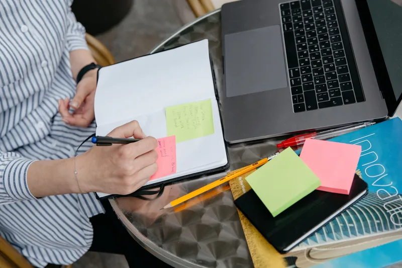 A person writing in a notebook next to a laptop and sticky notes on an organized desk