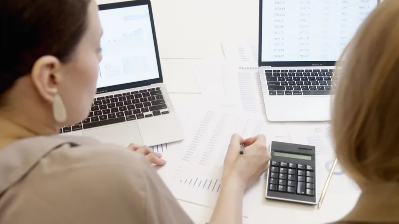 Two people reviewing spreadsheets and calculating numbers on laptops
