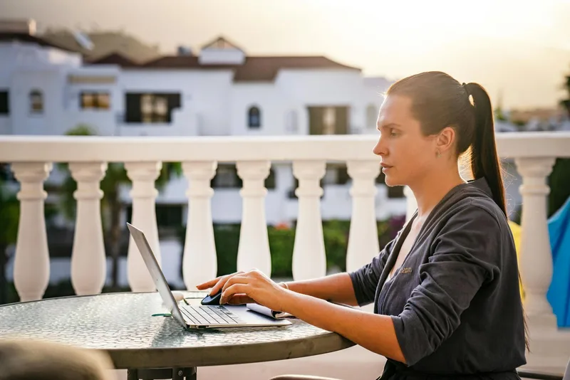 Woman working on laptop on a sunny balcony, independent and focused — representing the flexibility of remote VA work